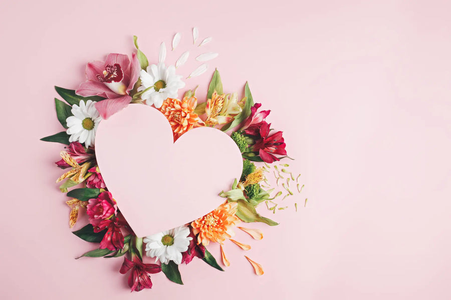 Herz aus Papier umgeben von bunten Blumen auf rosa Hintergrund.